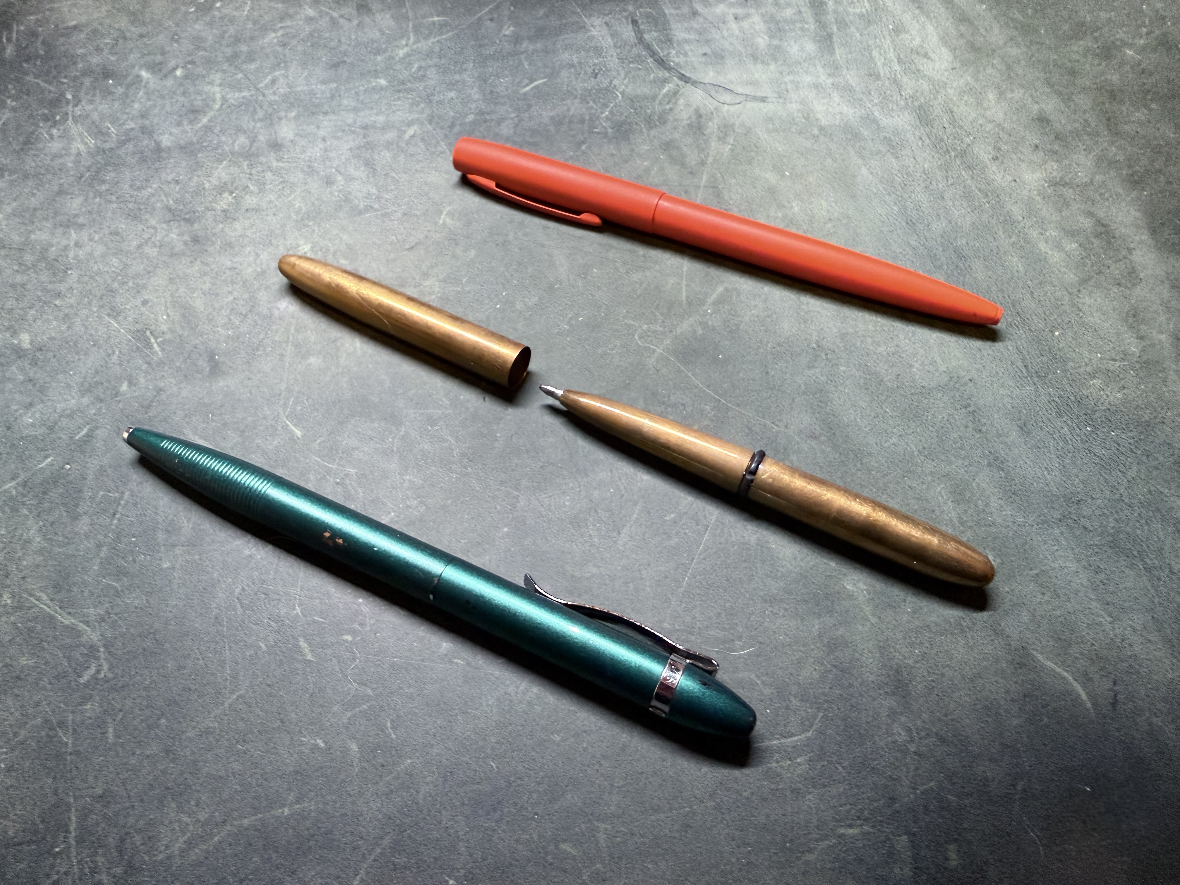 Three Fisher Space Pens: a green satin Vortex, an uncapped raw brass bullet and a hi-viz orange Cerakote Cap-O-Matic